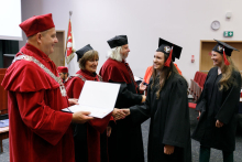 A man (pro-rector) in a red toga presents diplomas to graduates.