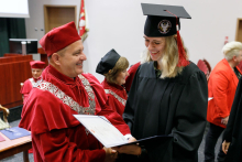 A man (pro-rector) in a red toga presents diplomas to graduates.