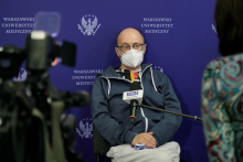 A man wearing glasses and a medical mask, dressed in a sweatshirt and sweatpants sits on a chair. Behind him is a wall in navy blue with the WUM logo. In front of him are television cameras. The man holds a microphone in his hand.