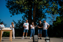 Six people stand on the volleyball court as if on a stage. Five of them are wearing T-shirts that say "2023 Chłapowo." A girl with long black hair speaks into a microphone.