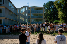 In the foreground, four people - three facing backwards, one sideways - speak into a microphone. A volleyball court is visible. Behind it, dozens of young people. 