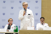 Three people, two sitting at a conference table, one standing - a man and holding a microphone in his hand. All are dressed in white medical gowns. Behind them is a blue wall with the WUM logo. 