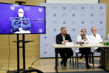 A group of seated people, two men and a woman. Two people are dressed in white doctor's aprons, one in a suit and has a tie. Next to them on a stand is a television on which a man is displayed. Behind them is a wall in blue with the WUM logo.