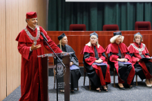 Graduation ceremony. In the foreground is a man (pro-rector) in a red toga at the lectern. Seated in the back are four people also in togas.