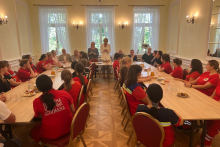 Women in red costumes sit at tables arranged in a "U" shape. Two women stand one speaks, the other translates her words.