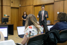 A man in a suit and glasses. He stands in front of the audience. He gestures.