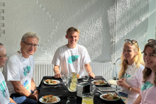 Five people wearing T-shirts with the inscription "2023 Chłapowo" sit at a table. On the table plates with snacks.