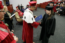 A man in a red toga hands a diploma to a woman in a black toga.