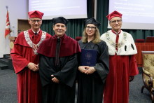 Four people in togas pose for a photo. Two men in red, one in black and one woman in black.