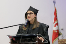 A woman in a black toga at the lectern, speaks into the microphone.