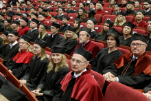 Auditorium. Many people in togas and birettas sit on chairs