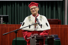 A man in a red toga and red biretta at the lectern, speaks into the microphone.