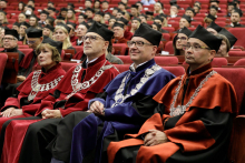 Auditorium. Many people in togas and birettas sit on chairs, In the foreground four deans.