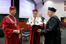 Two men in red togas hand a diploma to a man in a black toga.