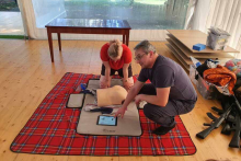 A blanket is spread on the floor. On the blanket, a young woman is practicing chest compressions on a mannequin. An older man is watching.