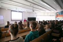 Sala wykładowa wypełniona słuchaczami. W oddali postać prelegentki. Za nią ekran.