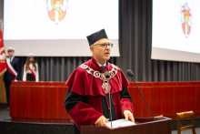 A middle-aged man in a red and black toga, glasses and academic cap speaks into the microphone. 