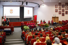 A projection of a large room with red chairs, people are sitting on chairs, a man is standing in front of them, behind a lectern, next to them, several people are sitting on chairs, behind them a multimedia screen with the department logo in maroon.