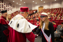 A middle-aged man dressed in a rector's toga shakes hands with a young girl, dressed elegantly, with a red and white sash and an academic cap.
