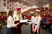 Mężczyzna w średnim wieku ubrany w togę rektorską ściska dłoń młodej dziewczynie, ubranej elegancko, z czerwono-białą szarfą i czapką akademicką. 