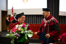 On the left, a woman dressed in an academic toga, holding a bouquet of flowers. On the right, a middle-aged man wearing an academic toga. 