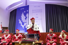 A middle-aged man stands behind the lectern. He is dressed in a rector's toga in red and white. In his right hand he holds a microphone. Behind him sit several people dressed in academic togas. In the background is a grey curtain with a white and blue decoration on it.