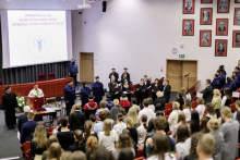 Rzut z góry na salę, w rzędach z czerwonymi fotelami mnóstwo osób, na środku stoją ludzie w togach akademickich, za nimi ekran multimedialny z niebieskim logo wydziału.