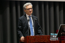 A middle-aged man, dressed in a suit and blue tie and wearing glasses stands behind the lectern and speaks