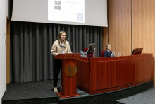 A woman wearing a light-coloured blouse stands behind a lectern and speaks, a multimedia screen in the background, two people sit next to her