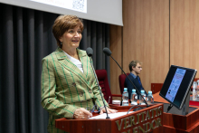 A woman in a green jacket, stands behind the lectern and speaks. Next to her sits a person. 