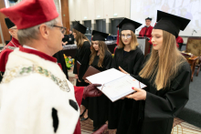 Grupa osób ubranych w czarne togi akademickie oraz mężczyzna ubrany w togę białą-czerwoną, który wręcza jednej osobie otwarty dyplom w okładce.