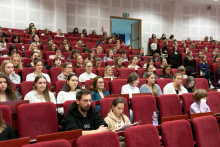 A group of young people sit in a lecture hall on red chairs
