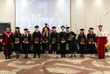 Grupa młodych osób ubranych w czarne togi akademickie stoi z dyplomami w rękach, uśmiecha się do aparatu. Obok rektor ubrany w czerwono-białą togę akademicką.