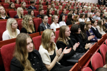 Young people dressed in gala attire sit on red chairs and applaud.