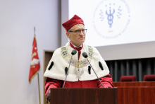 A man in a red and white toga, glasses and red academic cap speaks into a microphone. Behind him, a multimedia screen with a navy blue faculty logo.