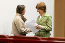 Two women - on the left wearing a light-coloured blouse, on the right wearing a green blazer - shake hands and smile.