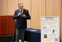 A middle-aged man in a suit and tie stands. In his right hand he holds a microphone raised to his mouth. In front of him is a poster of a conference