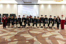 Grupa młodych osób ubranych w czarne togi akademickie stoi z dyplomami w rękach, uśmiecha się do aparatu. Obok rektor ubrany w czerwono-białą togę akademicką oraz dziekan ubrany w togę czarno-czerwoną.