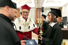 Trzy osoby, dwóch mężczyzn oraz młoda kobieta. Mężczyźni podają kobiecie dyplom oraz nagrodę, uśmiechają się.