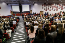 A projection of the hall from above, a large group of young people stand upright in rows with chairs. In front of them, in the middle of the room stands a group of people dressed in academic togas. Behind them a multimedia screen with a blue faculty logo.