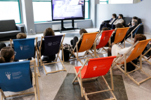 Colourful deckchairs spread out in the corridor, young people sitting on them. A screen is set up in front of them, displaying the broadcast of the inauguration.