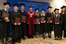 Grupa młodych osób ubranych w czarne stroje akademickie pozuje do zdjęcia z dyplomami w brązowych okładkach. W środku, między młodymi, stoi dziekan, ubrany w czaro-czerwoną togę.