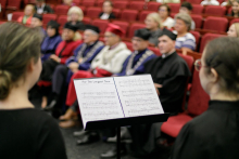 Pulpit muzyczny z nutami utworu. Tyłem do aparatu - członkowie chóru ubrani na czarno, przodem - osoby ubrane w togi akademickie.