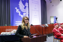 A middle-aged woman, with longer hair and wearing glasses, stands behind the lectern. Behind her, an inauguration decoration in white and blue with an eagle. 