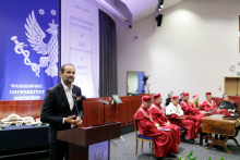 Mężczyzna w średnim wieku, w garniturze i białej koszuli stoi za mównicą. Za nią dekoracja inauguracyjna w biało-niebieskim kolorze z orłem. 