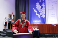 Mężczyzna w czerwonej todze akademickiej, z łańcuchem na szyi stoi za mównicą i przemawia. Za nim widać dekorację inauguracyjną w kolorach białym i niebieskim oraz poczet sztandarowy.