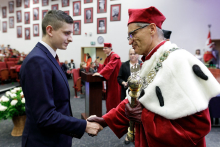 Mężczyzna w średnim wieku ubrany w togę w kolorze czerwonym i białym gratuluje młodemu mężczyźnie ubranemu w garnitur. W tle inne osoby.