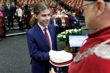 A middle-aged man wearing a toga in red and white congratulates a young man dressed in a suit. Other people in the background.