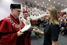 A young woman with fair hair on the right receives an academic cap from the hands of a middle-aged man - on the left. A man wearing a toga in black and red. Behind him a second man in a toga. In the background, a hall filled with people.