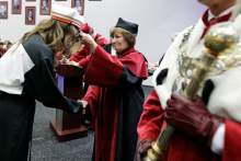 A middle-aged woman in a red and black academic toga places a cap on a young girl's head - a symbol of matriculation.